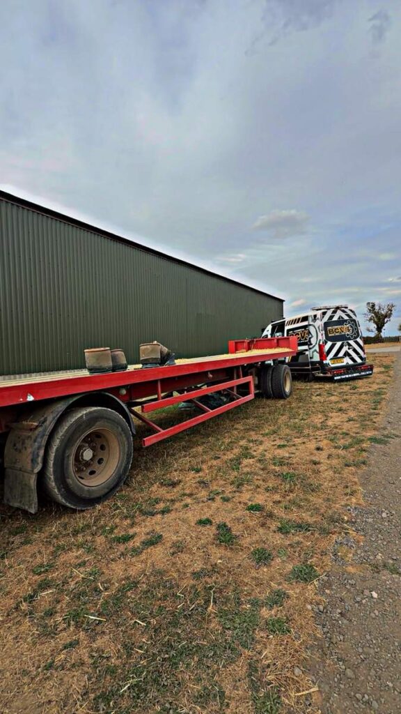 Red flatbed agricultural trailer parked outside a farm building awaiting repair