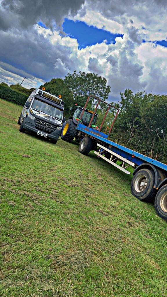 BCVS Mercedes Sprinter service van parked in a grass field beside a green tractor and a blue flatbed bale trailer