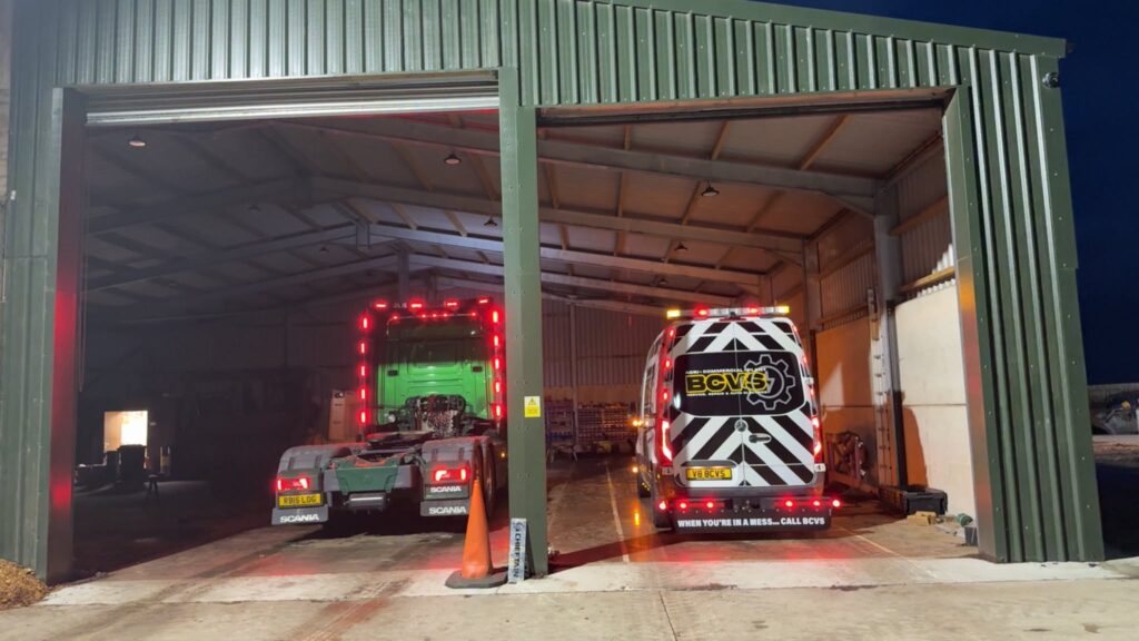 BCVS van and a green Scania tractor unit parked inside a lit workshop shed after dark