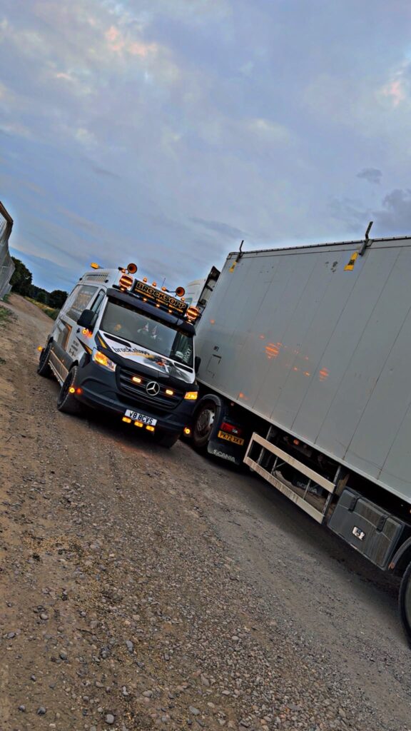 BCVS van with hazard lights on, parked nose to nose with a Scania box trailer on a gravel yard at dusk